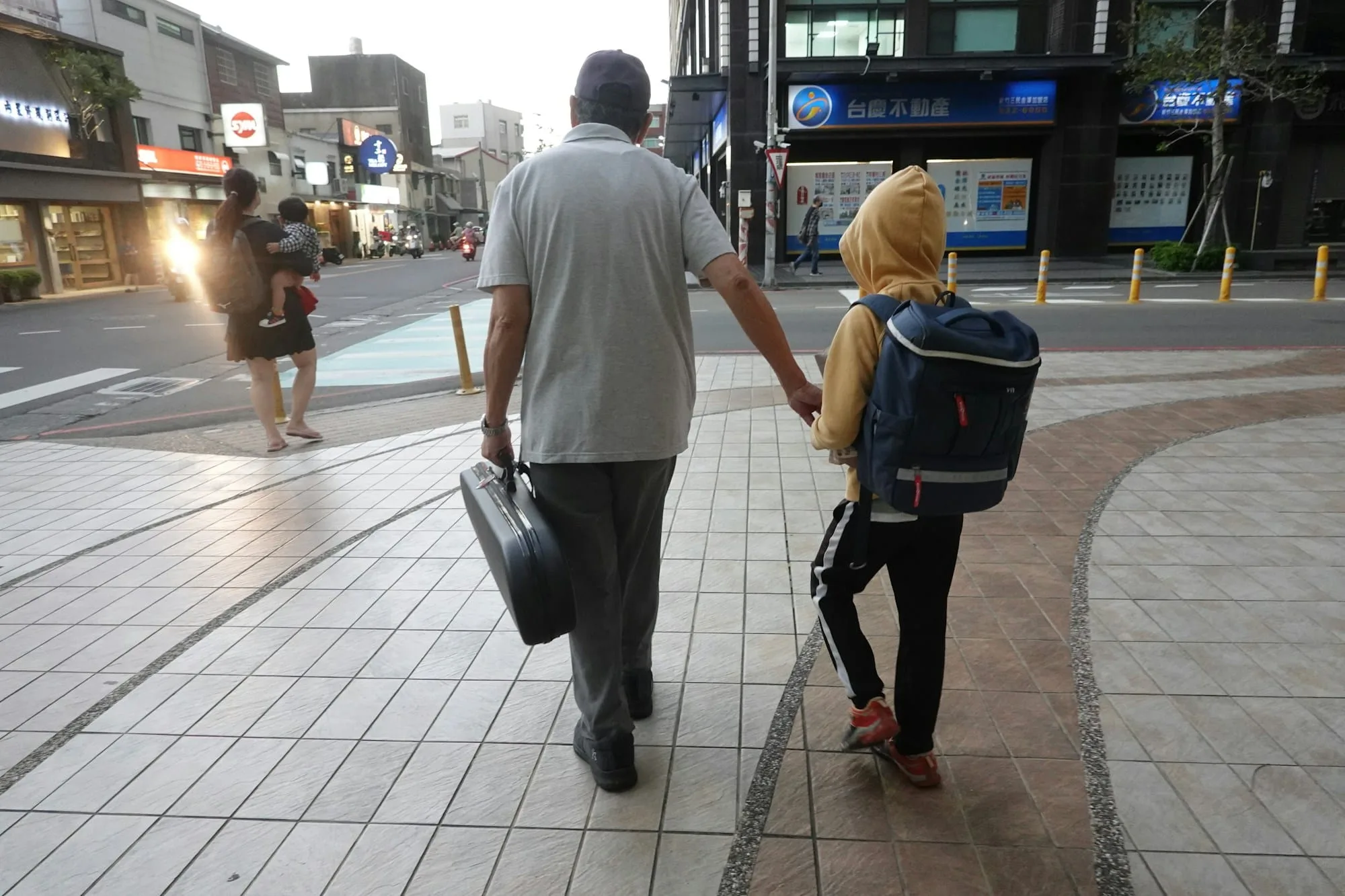 An adult and a child with a backpack walking hand in hand along a city pavement, seen from behind