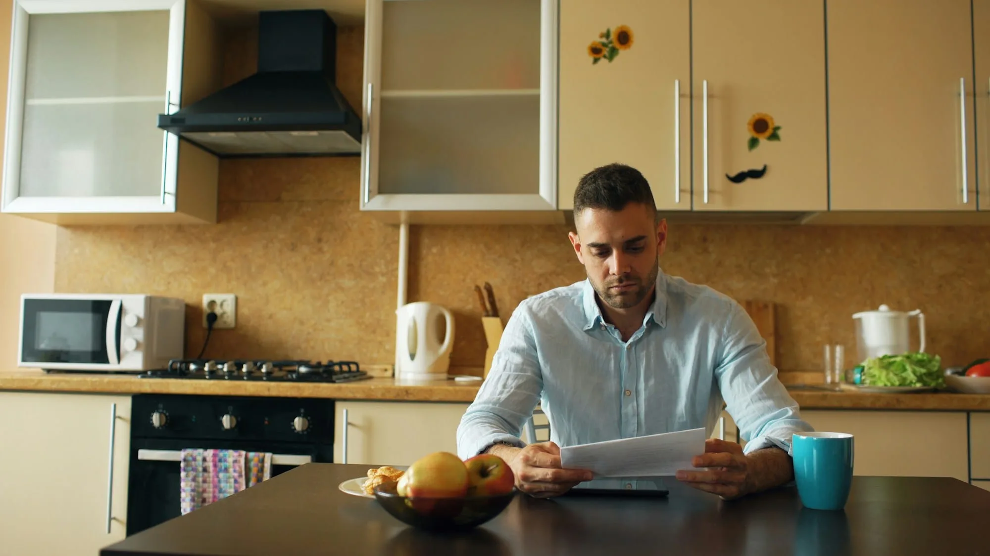 A man sitting at a kitchen table reading a letter with a serious expression